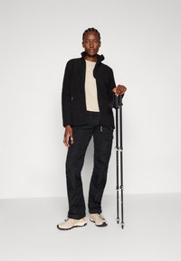 Fleece jacket in black, beige top, black trousers with pockets, and beige shoes. Holding trekking poles, standing against a light grey backdrop.
