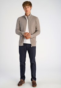 Beige stickad ziptröja med ståkrage, i kombination med en vit t-shirt, marinblå byxor och bruna segelskor. Enkel design med texturerad tyg.