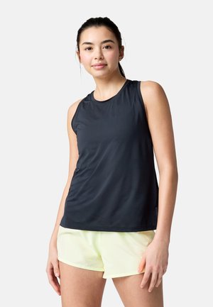 Giovane donna con top sportivo nero senza maniche e pantaloncini giallo chiaro, in piedi davanti a uno sfondo chiaro e uniforme.