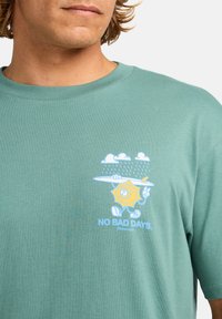 Mentazöld pamut póló, amelyen egy mosolygó nap grafikája látható, amely mellett egy szörfdeszka, felhők és a kék színű "NINCS ROSZ NAP" felirat található.