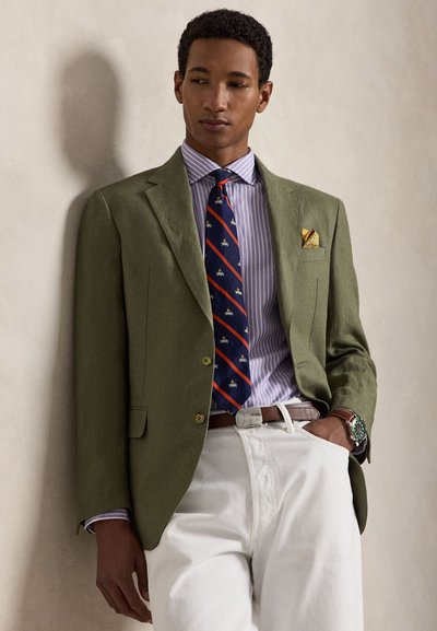 Άνδρας γέρνει σε τοίχο φορώντας ελιά πράσινο σακάκι, ριγέ μωβ πουκάμισο, μοτίβο σκούρα μπλε γραβάτα, λευκό παντελόνι και καφέ ζώνη και ρολόι.