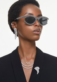 Óculos de sol preto em forma de olho de gato com detalhes em cristal, brincos longos prateados, colares de prata em camadas e um broche decorativo num blazer escuro.