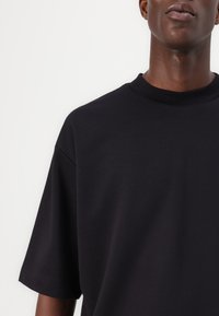 Μαύρο βαμβακερό μπλουζάκι με χαλαρή εφαρμογή, στρογγυλή λαιμόκοψη και κοντά μανίκια. Απαλή υφή και μινιμαλιστικός σχεδιασμός, χωρίς ορατά μοτίβα.