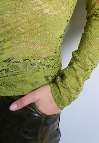 Main dans la poche portant un haut en dentelle florale vert olive et un pantalon en cuir marron foncé sur un fond uni.