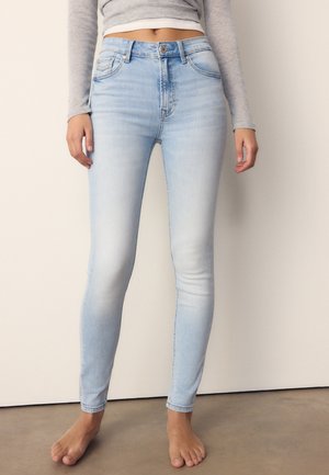 Kvinde iført højtaljede, lyseblå skinny jeans og en grå, langærmet crop top, stående barfodet på en grå gulv.
