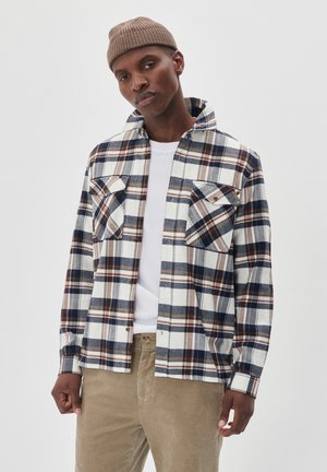 Plaid flannel shirt in cream, navy, brown, and gray. Features two chest pockets, button-down collar, and textured fabric. Worn over a white shirt.
