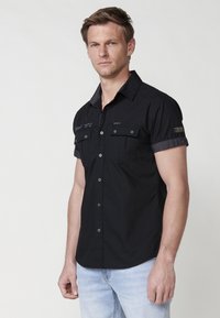 Camisa de botones de manga corta negra con dos bolsillos frontales, cuello en contraste y costuras decorativas. Textura suave, corte casual.