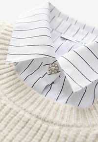 Beige strikket sweaterkrave med en hvid skjorte med sorte lodrette striber. Dekorativ klar perleknapdetalje på skjorteskraven.