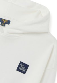Witte kinderhoodie met een trekkoordkap. Voorzien van een marineblauw vierkant label met de tekst "IK BEN BEPERKT EDITIE". Zachte stoftextuur.