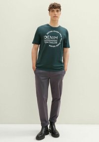 Camiseta de algodón verde con texto gráfico blanco, combinada con pantalones gris y botas negras hasta el tobillo. Diseño sencillo, ajuste casual.