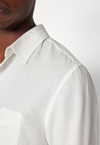 Camicia bianca in lino con colletto classico, taschino sul petto e una trama delicata, che mostra un tessuto morbido e un design affusolato.