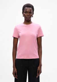 Pink bomulds t-shirt med rund halsudskæring og korte ærmer, med et lille logo centreret under halsen. Bæres med sorte bukser.