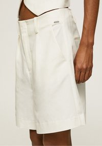 White linen shorts featuring a relaxed fit, front pleats, and side pockets. Includes a small metallic brand tag on the waistband.