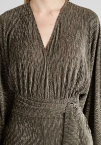 Vestido envolvente gris texturizado con escote en V, que presenta detalles fruncidos y un cinturón atado en la cintura. Hilos que reflejan la luz crean un brillo sutil.