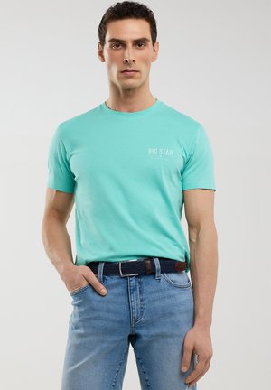 Gai turquoise kokvilnas t-krekls ar apaļu kaklu, īsām piedurknēm un "BIG STAR" logo. Saskaņots ar zilu džinsu biksēm un austiem tumši ziliem jostu.