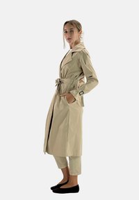Beige trenchcoat med strukturerad silhuett, vid krage, midjebälte och två framfickor, tillsammans med ljusa byxor och svarta ballerinaskor.