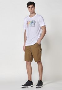 T-shirt bianco in cotone con grafica di bicicletta multicolore, abbinato a pantaloni cargo marroni e scarpe da ginnastica nere, su uno sfondo neutro.