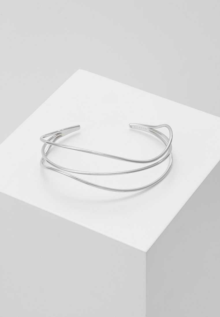 Bracelet manchette minimaliste en argent avec trois bandes incurvées superposées, présenté sur un socle blanc sur fond gris.