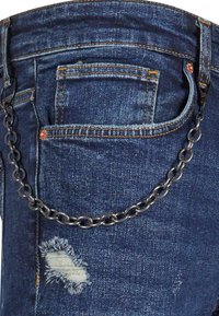 Jeans de denim desgastado en azul oscuro con un parche deshilachado, que cuentan con una cadena de metal plateado adjunta en el bolsillo trasero para un detalle adicional.
