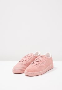 Paire de baskets en daim rose avec lacets et doublure intérieure blanche sur une surface et un fond blancs.