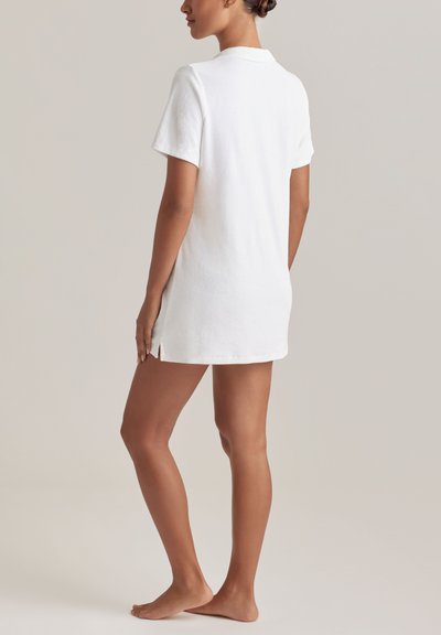 Γυναίκα όρθια, ξυπόλυτη, φορώντας λευκό φόρεμα από περκάλι με κοντά μανίκια και πλαϊνές σχισμές, εμφανίζεται από πίσω σε απλό φόντο.