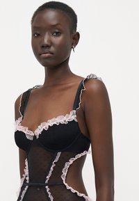 Lingerie nera con pannelli trasparenti, rifinita con morbidi volant rosa lungo i bordi. Presenta una texture a pois e spalline a contrasto.