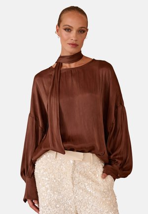 Femme portant un chemisier marron brillant à manches longues avec un foulard noué autour du cou et un pantalon crème texturé taille haute sur un fond uni.