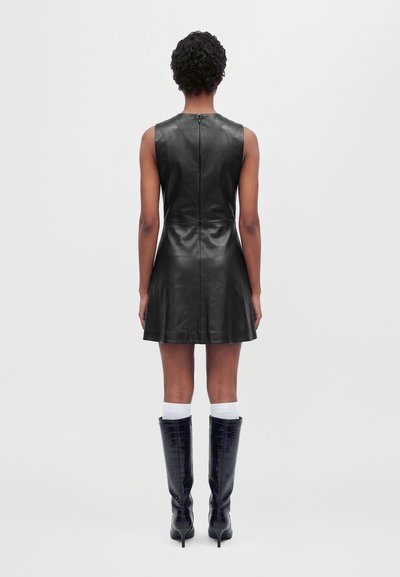 Mannequin vu de dos, portant une mini-robe noire en cuir sans manches, des chaussettes blanches et de hautes bottes noires à talons, sur un fond uni.