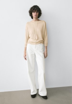 Kvinde med krøllet hår iført beige langærmet top, langt vedhængshalskæde, brede hvide bukser og sorte sko, stående op ad en hvid væg.