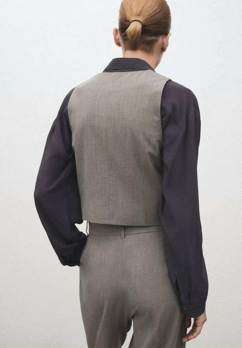 Chaleco gris corto hecho de tejido texturizado, combinado con una camisa negra de manga larga. Incluye cuello con solapa y cintura ajustada.