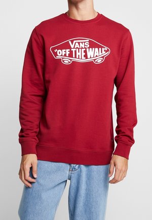 Osoba nosząca czerwoną bluzę Vans z długim rękawem z białym logo deskorolki "Off The Wall", w połączeniu z jasnoniebieskimi dżinsami.