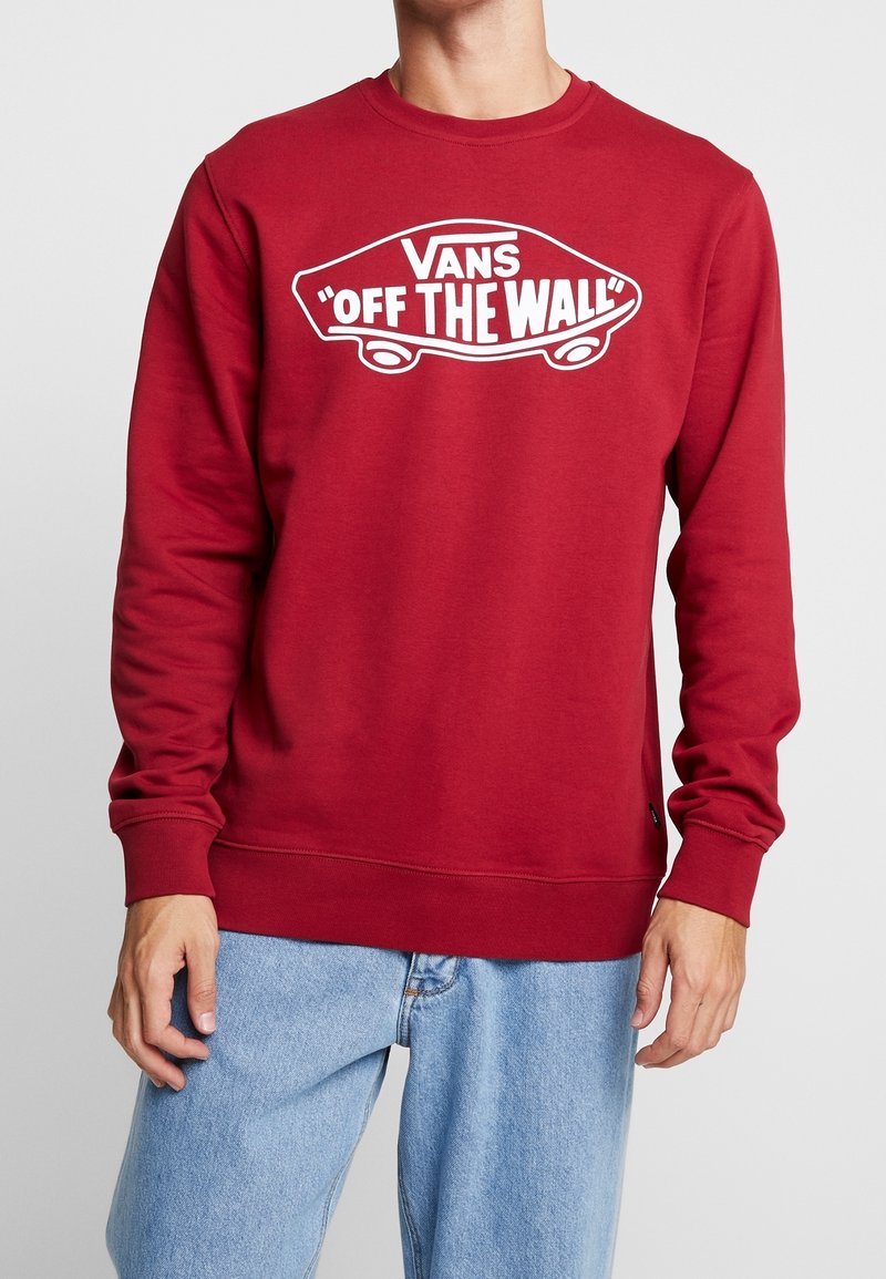 Personne portant un sweat-shirt Vans rouge à manches longues avec le logo skateboard blanc "Off The Wall", associé à un jean bleu clair.