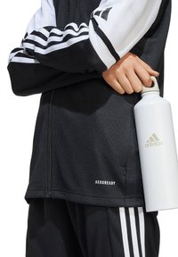 Schwarze Zip-Jacke mit weißen Streifen und "AEROREADY"-Logo am Saum; Hand hält eine weiße Wasserflasche mit dem Adidas-Logo.