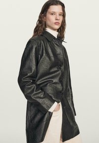 Černý kožený kabát s texturovaným, vzorovaným povrchem, se zapínáním na knoflíky, límcem a bočními kapsami. Nenošený přes lehký svetr.