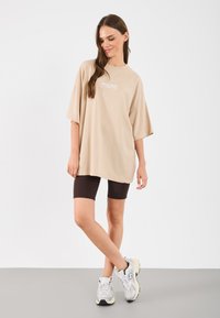 T-shirt oversize bege de mangas curtas com detalhe de logótipo, combinada com calções de bicicleta castanhos e sapatilhas brancas com detalhes cinzentos.