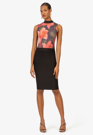 Frau trägt ärmelloses Kleid mit schwarzem Rock und Blumendruckoberteil, steht barfuß vor weißem Hintergrund.