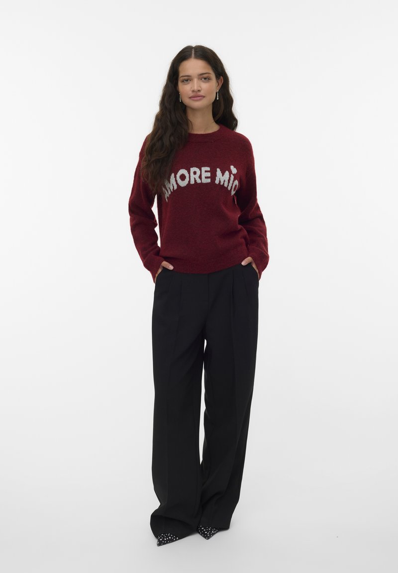 Roter Pullover mit "AMORE MIO" in weißen Buchstaben, kombiniert mit hochtaillierten schwarzen Hosen und spitzen Schuhen mit Nietenakzenten.