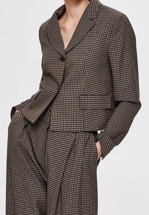 Kvinde iført en brun og sort hundetandsmønstret blazer og matchende plisserede bukser med hænderne i lommerne mod en ensfarvet baggrund.