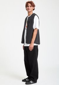 Grå fleecevest med en lynlås lomme, båret over en hvid graphic T-shirt. Sorte løse jeans og sorte sneakers med hvide striber.