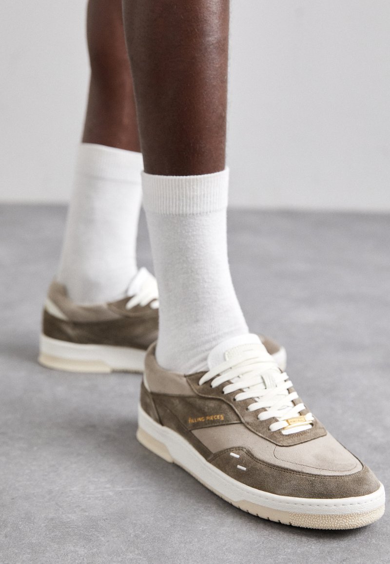 Baskets en daim marron avec des accents blancs, dotées de lacets, d'une semelle texturée et d'un détail en logo doré. Portées avec des chaussettes blanches à mi-mollet.