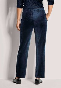 Vrouw van achteren in een donkerblauwe velours broek met wijde pijpen en zwarte glanzende loafers, staande tegen een lichte muur.