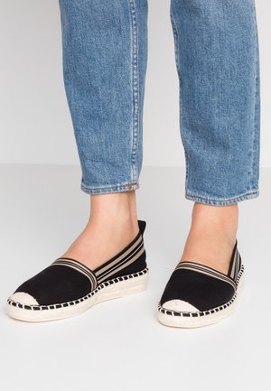 Person iført lyseblå jeans og sorte espadrille kiler med beige tåcapper og stribede elastikbånd.