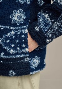 Marineblauwe fleecjas met witte bloemenpatronen, zachte textuur en een zijzak; beschikt over geribbelde manchetten en zoomdetails.