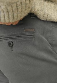 Pantalones grises hechos de una tela texturizada, con un bolsillo lateral, cierre de botón y una etiqueta de logo marrón. El diseño es simple y moderno.