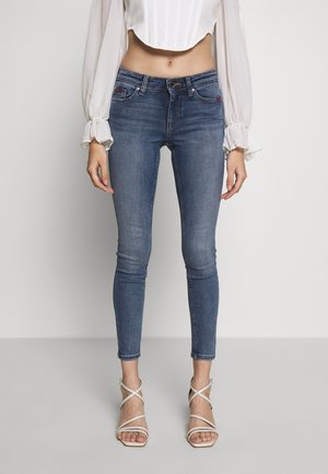 Femme portant un jean skinny bleu, un chemisier blanc à manches longues raccourci et des sandales blanches à talons hauts avec brides, debout devant un fond uni.