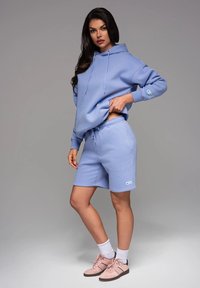 Felpa leggera color blu chiaro con cappuccio e lacci, pantaloni corti abbinati e un piccolo logo. Indossata con sneakers rosa pastello e calzini bianchi sopra la caviglia.