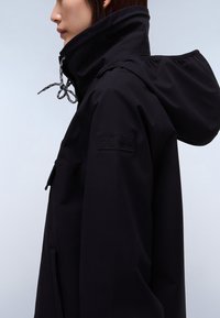 Persona con chaqueta negra de cuello alto con capucha ajustable y bolsillo en el pecho, mirando hacia la izquierda sobre un fondo claro.