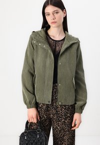 Maslinasto zelena kordura bomber jakna s kapuljačom, zatvaranjem na zip i gumbima. Uparena s crnim topom i hlačama s leopard uzorkom. Crna torba.