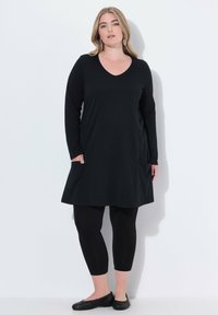 Schwarze Tunika mit langen Ärmeln und V-Ausschnitt, ausgestattet mit zwei seitlichen Taschen, kombiniert mit schwarzen Leggings und schwarzen flachen Schuhen. Glatte Stofftextur.