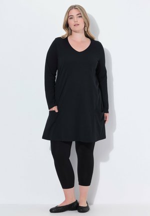 Tunika nera a maniche lunghe con scollo a V, dotata di due tasche laterali, abbinata a leggings neri e scarpe basse nere. Tessuto con texture liscia.